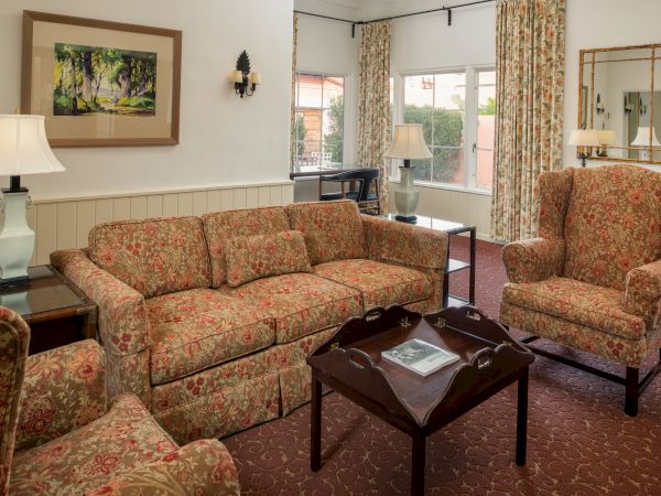 A cozy hotel living room with floral patterned sofa and chair, wooden coffee table, lamps, framed art, and a windowed backdrop.