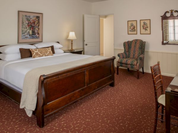 A classic hotel room with a wooden king bed, nightstand, dresser, comfy chair, desk, warm lamps, and traditional decor, cozy and inviting.