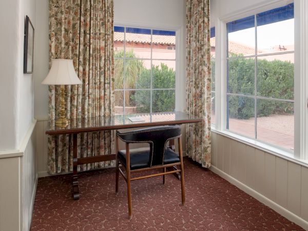 A small, sunlit office with a wooden desk and chair, floral curtains, a floor lamp, and large windows showing a sunny backyard.