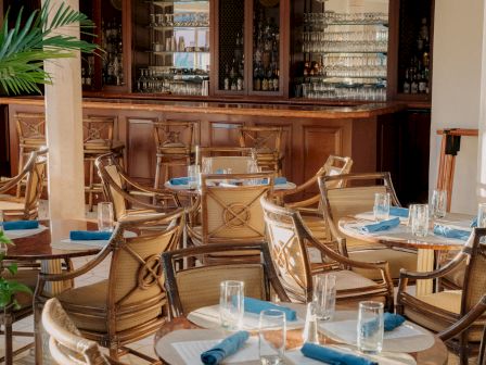 Cozy restaurant with wooden bar, wicker chairs, and neatly set tables; blue napkins, clear glasses, and plants add a welcoming touch.