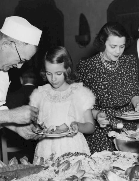 Chef in white hat serving cake to children at a party, while adults line up to choose desserts.