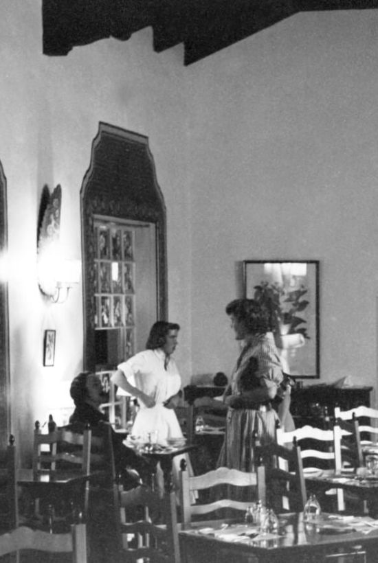 A retro dining room scene with two women standing and talking, several chairs around tables, and a framed mirror on the wall.