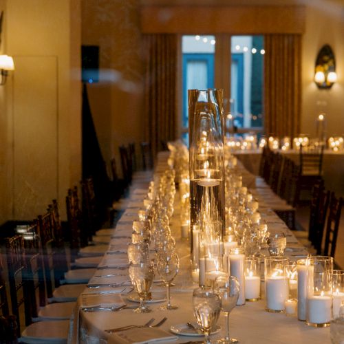 A long, elegantly set banquet table with candles, glassware, and chairs in a dimly lit room, ready for a formal dinner.