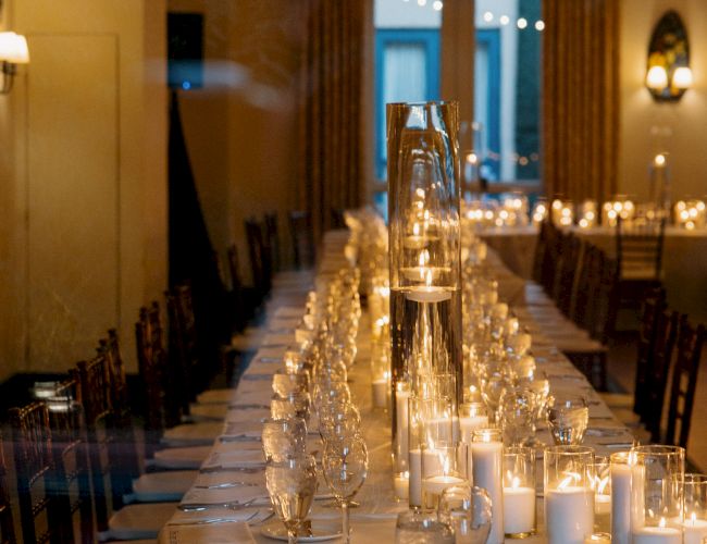 A long, elegantly set banquet table with candles, glassware, and chairs in a dimly lit room, ready for a formal dinner.