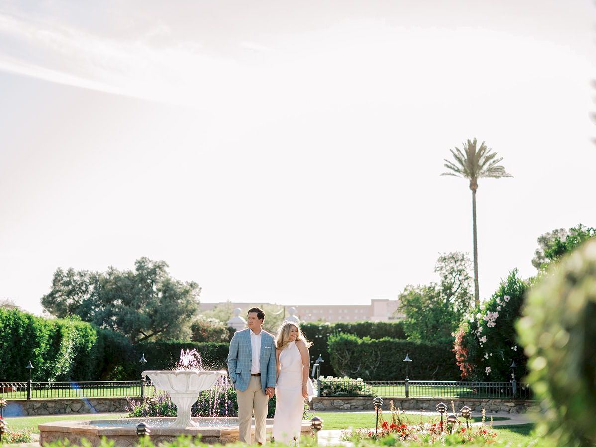 A couple walks hand-in-hand through a sunny garden with a fountain, lush hedges, and palm trees, enjoying a bright outdoor moment.
