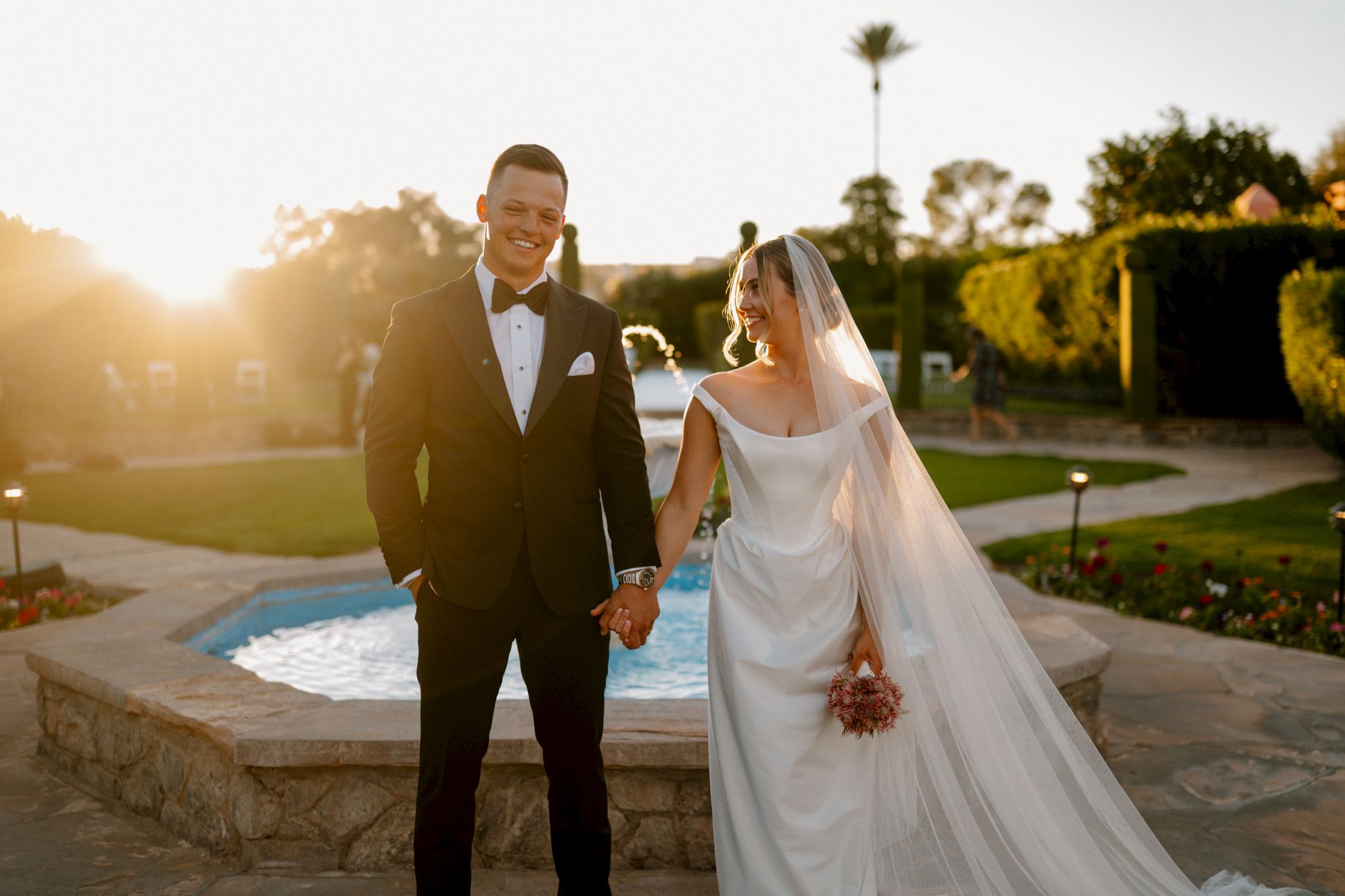 A newlywed couple walks hand-in-hand by a fountain at sunset; the groom in a black tux and the bride in a flowing white gown with a veil.