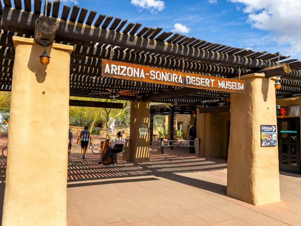 Entrance to the Arizona-Sonora Desert Museum, with a wooden sign and ornate shade pergola under a sunny blue sky.