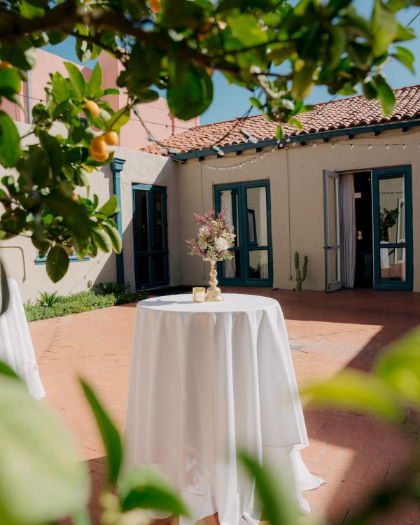A small round table with a white tablecloth and floral centerpiece sits outdoors near a Mediterranean-style house with open doors in the background.