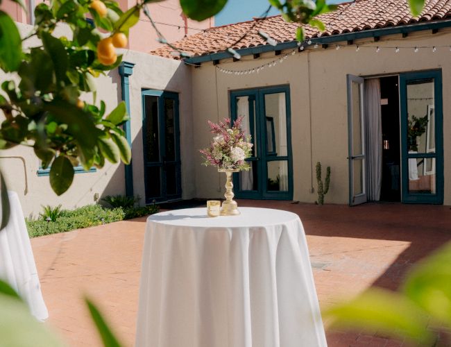 Sunlit courtyard patio with a white tablecloth, floral centerpiece, string lights, and blue-trimmed doors framed by lush greenery.