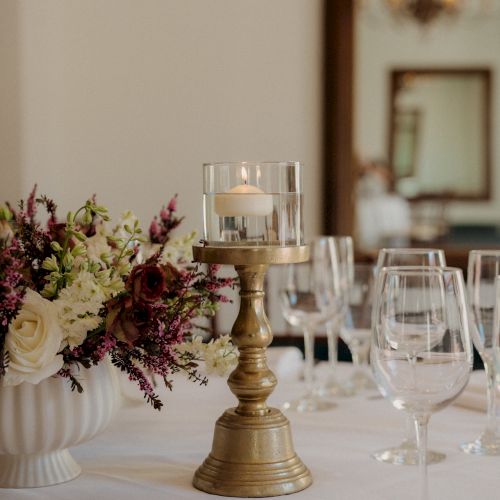 A centerpiece with a gold candle holder and glass candle, flanked by elegant flowers and multiple wine glasses on a white tablecloth.