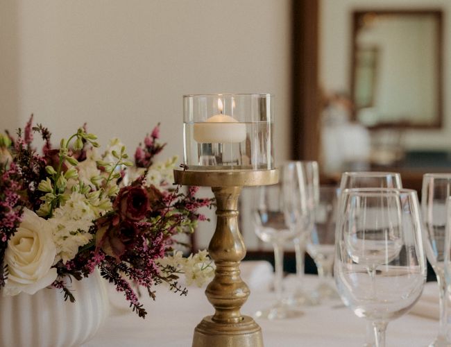 A centerpiece with a gold candle holder and glass candle, flanked by elegant flowers and multiple wine glasses on a white tablecloth.