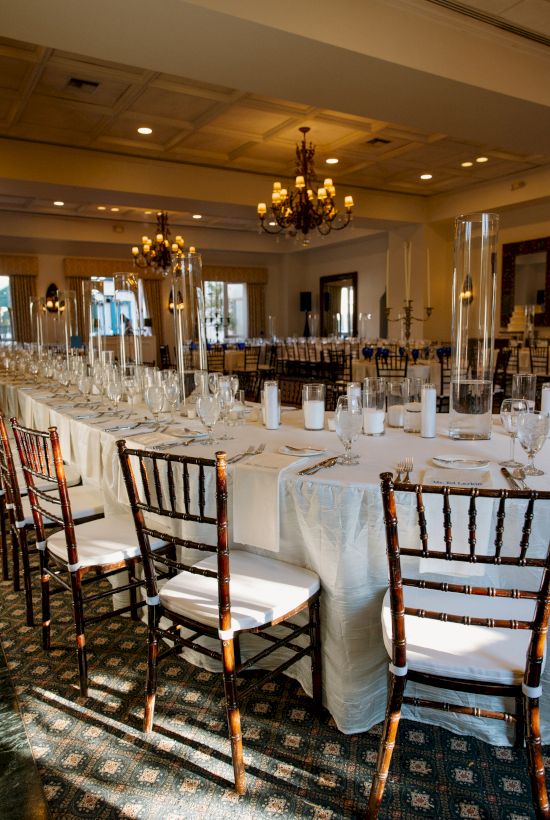 A formal dining setup with a long, pale tablecloth, elegant chairs, place settings, candles, and chandelier lighting in a banquet hall.