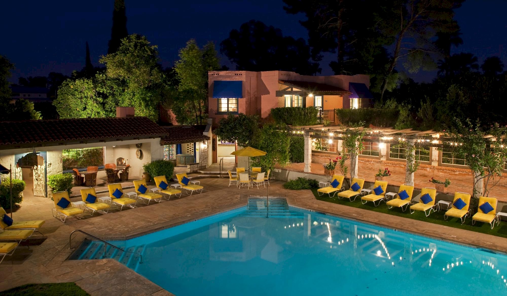 A lit outdoor hotel pool area at night with blue water, surrounding lounge chairs, palm trees, and a modern building backdrop.