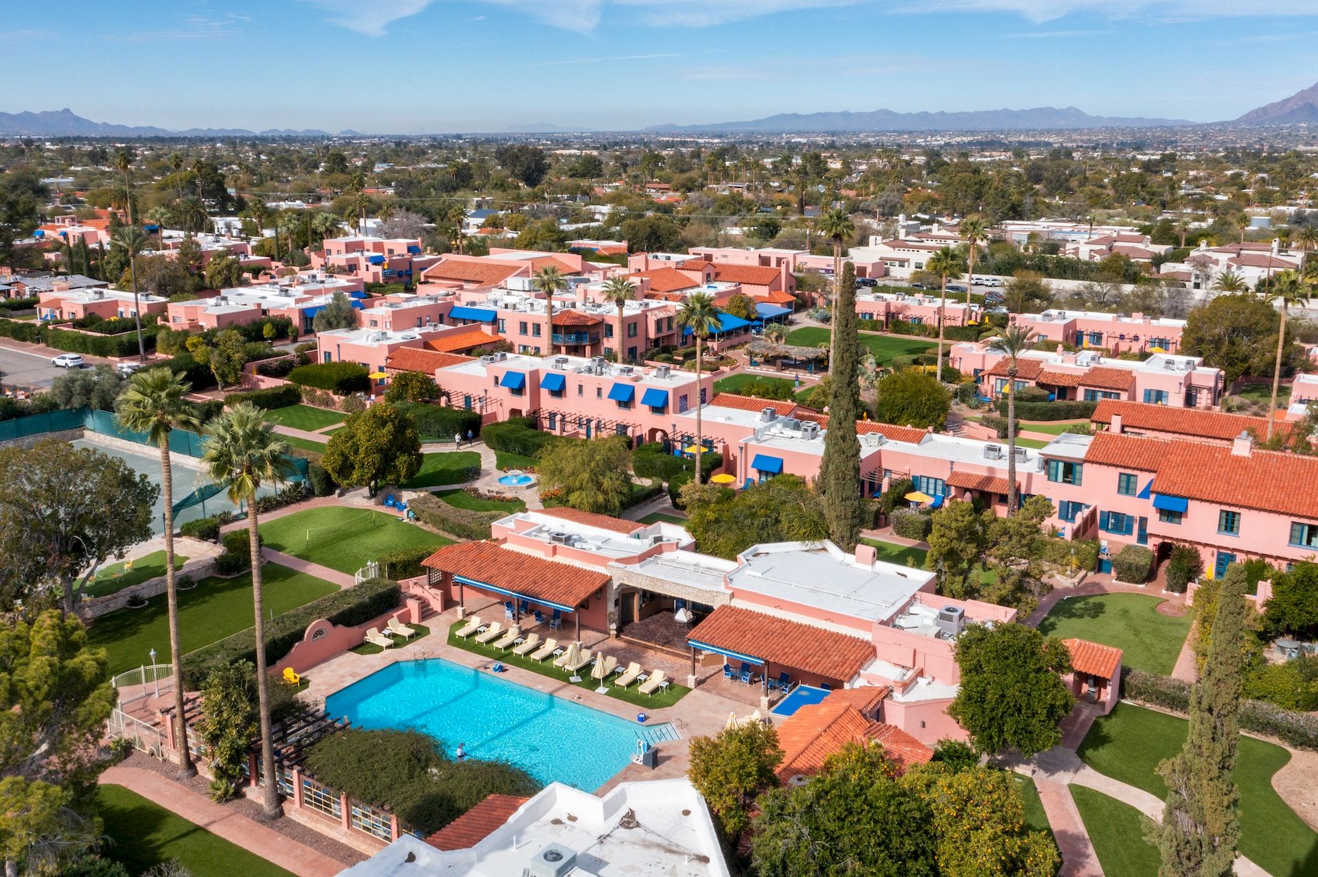 Aerial view of a sunny resort area with colorful houses, a central pool, palm trees, and manicured lawns, set against a distant desert landscape.