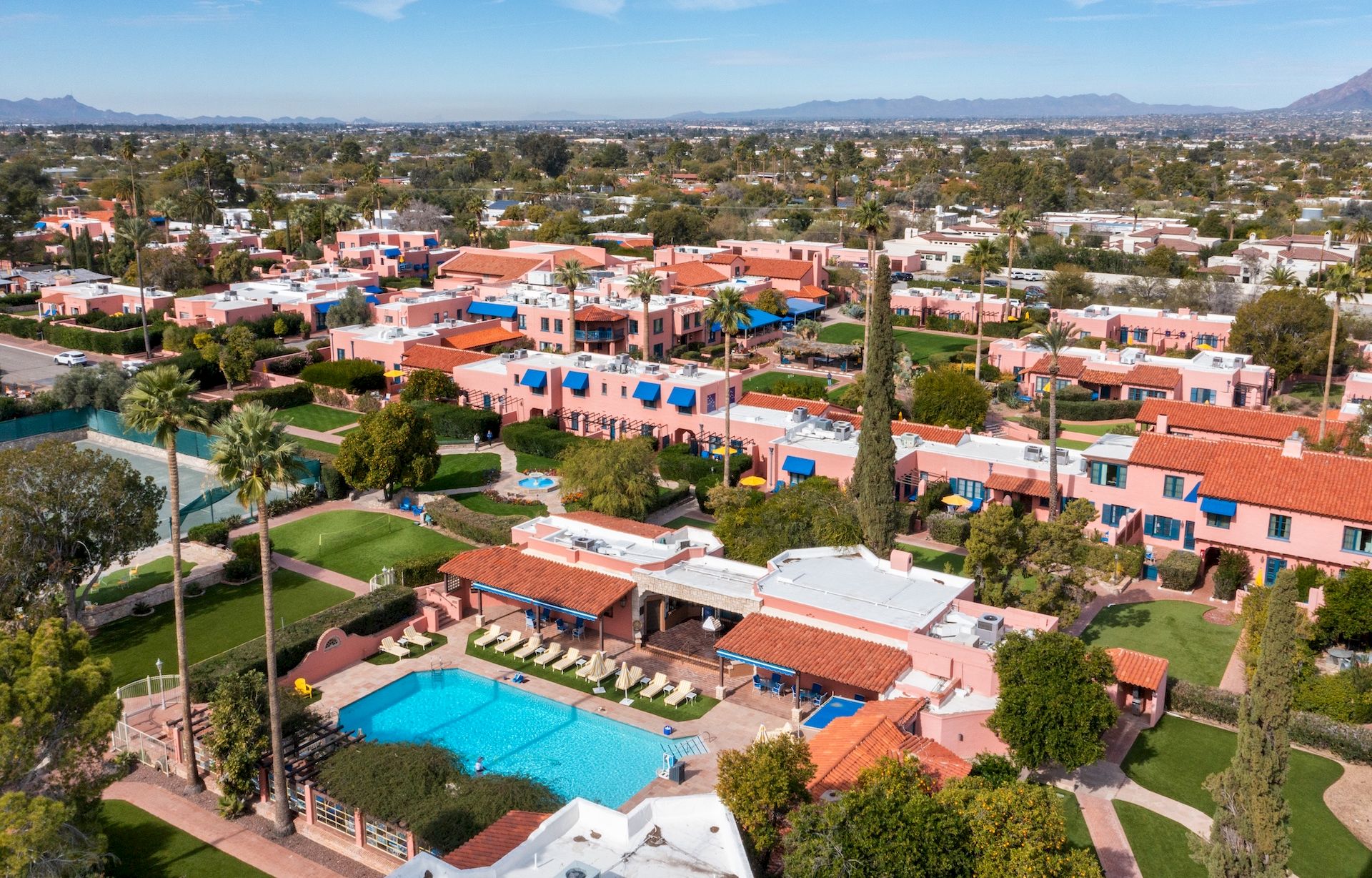 Aerial view of a sunny resort area with colorful houses, a central pool, palm trees, and manicured lawns, set against a distant desert landscape.