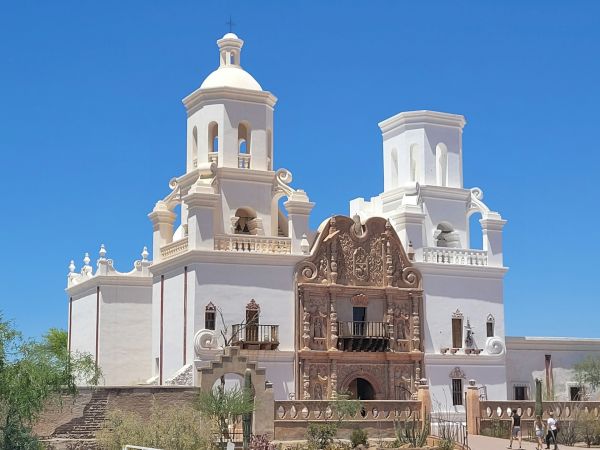 A white church with two towers and an ornate arched entrance, set against a bright blue sky, perched on a slight hill.