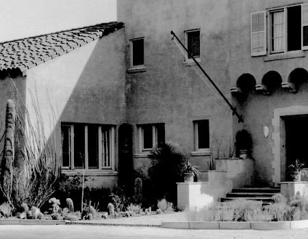 A grey stucco house with steps, arched windows, potted plants, and a small front yard on a sunny street.