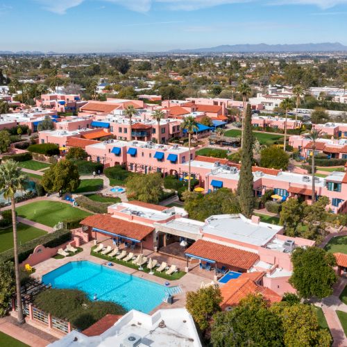 Aerial view of a resort with red-tiled roofs, a turquoise pool, palm trees, and sunlit buildings by clear blue water.