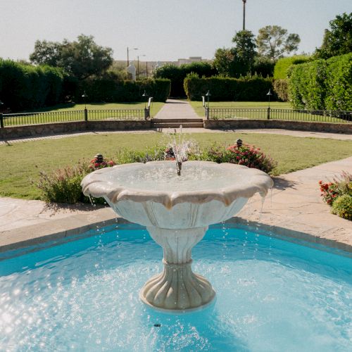 A decorative fountain in a sunlit garden with a sunny sky and manicured lawn behind it, centered by a tiered water feature.