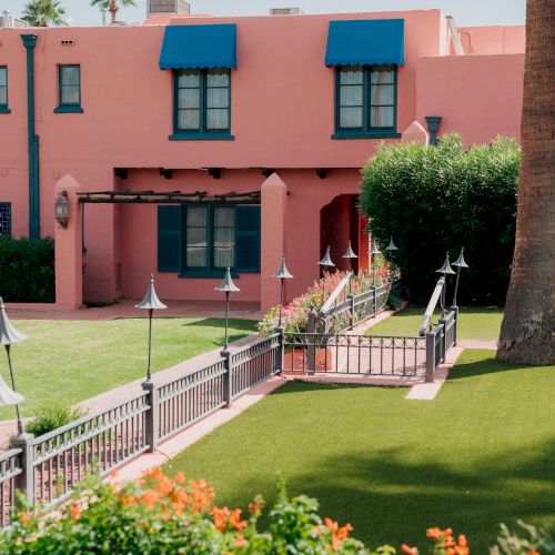 A colorful pink building with blue trim, arched windows, and a green lawn. A small white railing leads to a sunny path; cheerful garden blooms.