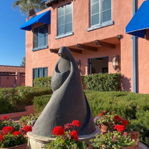 A small black tent or hooded structure in a vibrant garden with colorful flowers, beside peach-colored houses with blue shutters, under a clear sky.