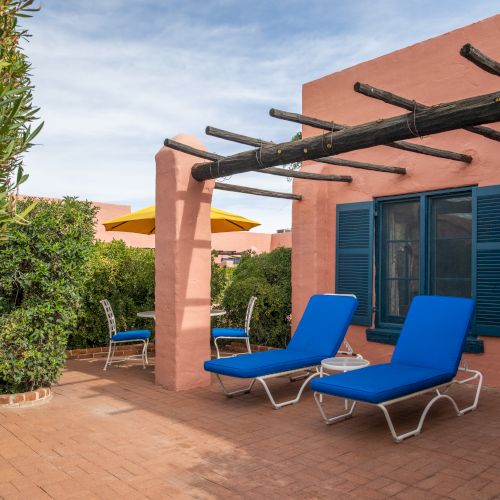 A sunny patio with pink stucco walls, blue lounge chairs, potted hedges, and a wooden pergola over a tiled deck.