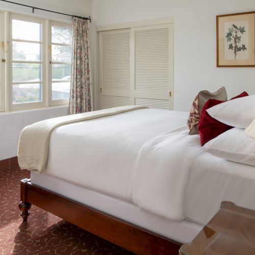 A cozy bedroom with a large bed dressed in white linens, wooden furniture, a bedside lamp, floral curtains, and a windowed wall&mdash;bright and inviting.