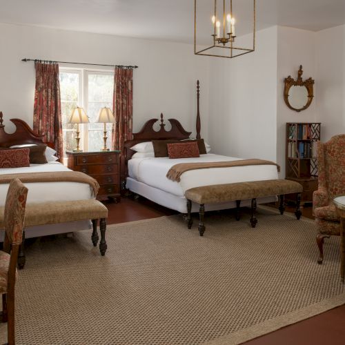 A spacious bedroom with two wooden four-poster beds, matching nightstands, a seating area, and a beige rug, giving a classic, vintage hotel vibe.