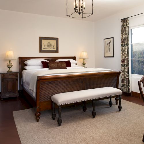 A traditional hotel room with a wooden king bed, matching nightstands, a bench at the foot, a sofa, and a small table by a window with floral curtains.