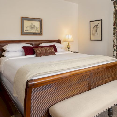 A cozy hotel bedroom with a wooden king bed, white linens, two nightstands, lamps, a bench at the foot, and patterned curtains&mdash;calm and classic.