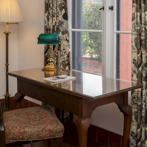 A classic wooden desk with a green banker&rsquo;s lamp, chair, and floral curtains by a window.