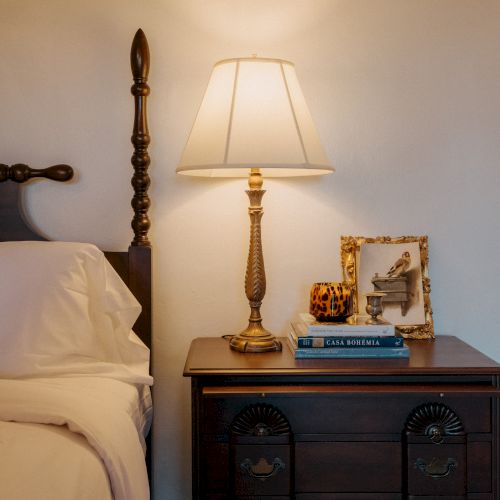A bedroom nightstand with a lamp, framed photo, and books beside a wooden bed post, softly lit by a warm table lamp at night.