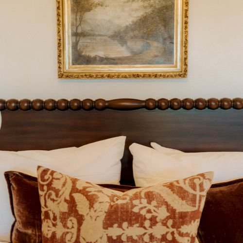 A neatly made bed with a carved wooden headboard, white pillows, and a decorative patterned throw pillow, framed landscape painting above the bed.