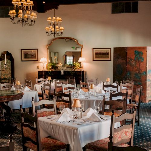 A cozy, classic dining room with white-draped tables, vintage chairs, chandeliers, and a warm, intimate ambiance for a refined meal.