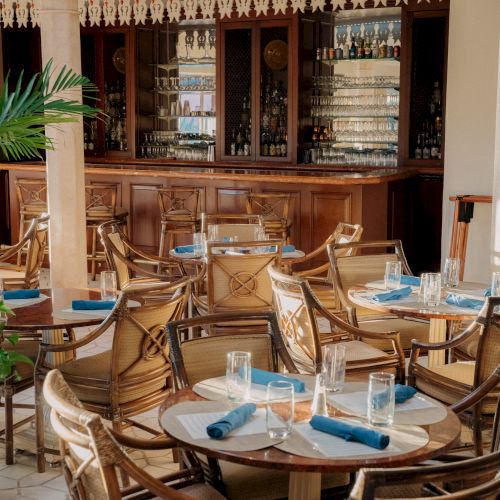 A cozy restaurant with round tables, blue napkins, glassware, and wooden chairs; a bar in the back showcases bottles and glass shelves.