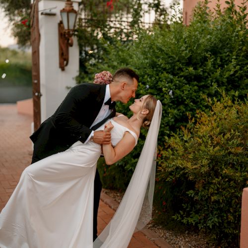 A newlywed couple shares a kiss on a sunlit path, he gently dips her in a romantic wedding pose.
