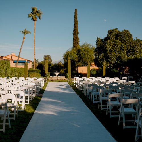An outdoor wedding aisle setup with rows of white chairs on either side, a white runner, palm trees, and sunny skies.