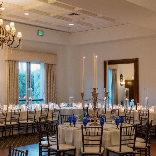 Elegant banquet setup in a bright room with round tables, white linens, and blue napkins; tall candlesticks and a large mirror add charm.