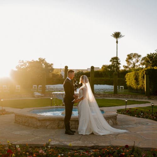 Two people in wedding attire stand by a fountain, holding hands and looking at each other as the warm sunset lights the garden.