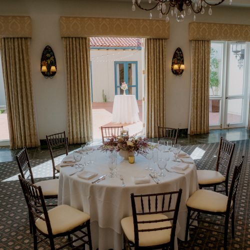 A formal dining room with a round table set for eight, white tablecloths, chandeliers, and large windows leading to an outdoor area, elegant decor.