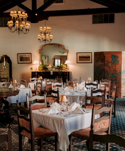 A cozy, upscale dining room with white-tablecloths, folded napkins, wooden chairs, warm lighting, and a decorative fireplace.