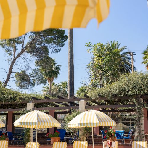 An outdoor resort pool with yellow-and-white striped umbrellas, lounge chairs, and a woman relaxing in sunny greenery.