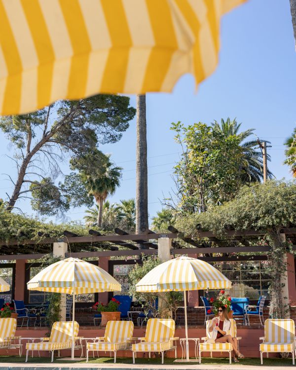An outdoor resort pool with yellow-and-white striped umbrellas, lounge chairs, and a woman relaxing in sunny greenery.