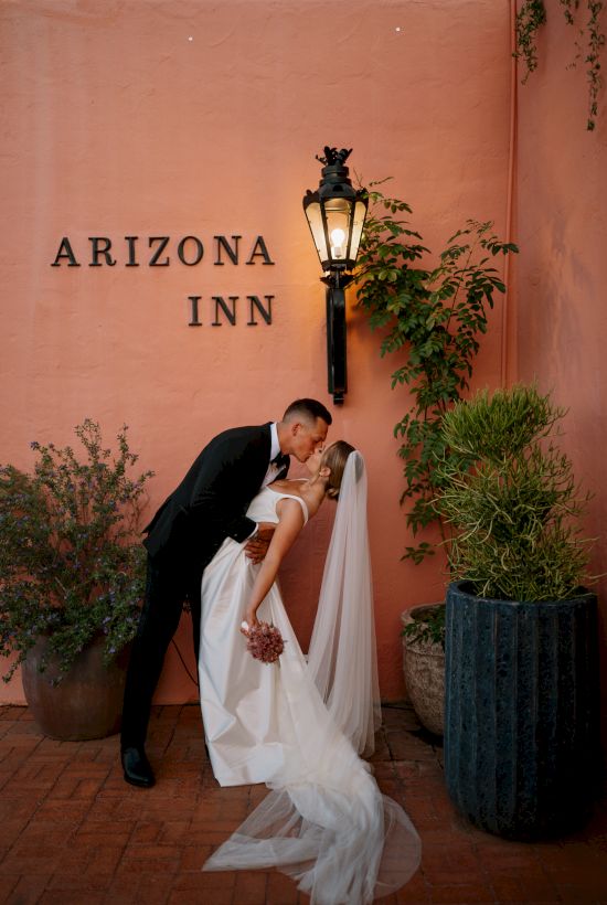 A newlywed couple share a kiss by an orange wall with &ldquo;ARIZONA INN,&rdquo; a hanging lantern, and potted plants, creating a romantic outdoor scene.
