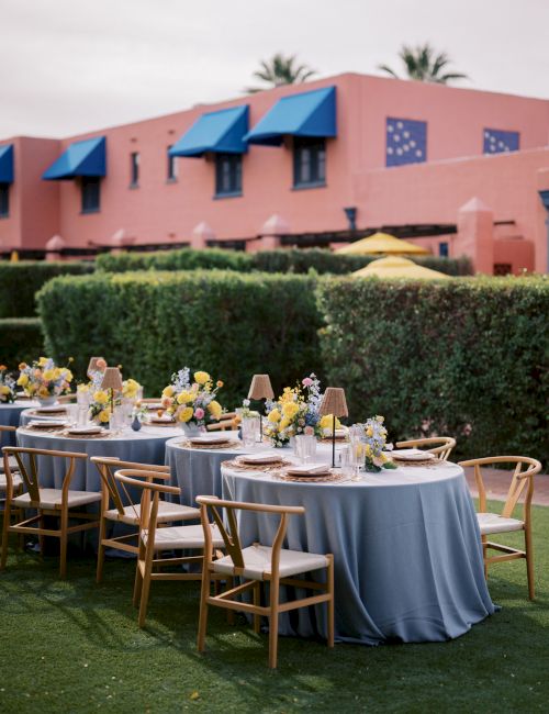 An outdoor banquet setup with round tables, floral centerpieces, chairs, and a red brick building with blue awnings.
