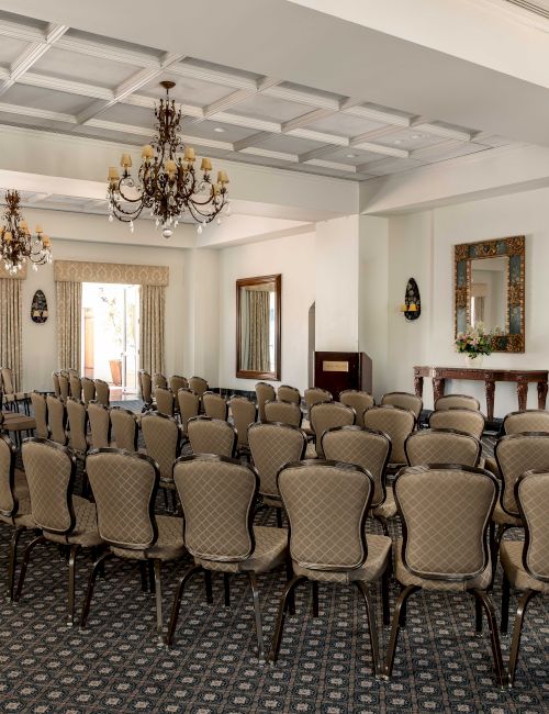 An elegant ceremony hall with rows of upholstered chairs, ornate chandeliers, tall curtains, and a decorated side table with floral display.