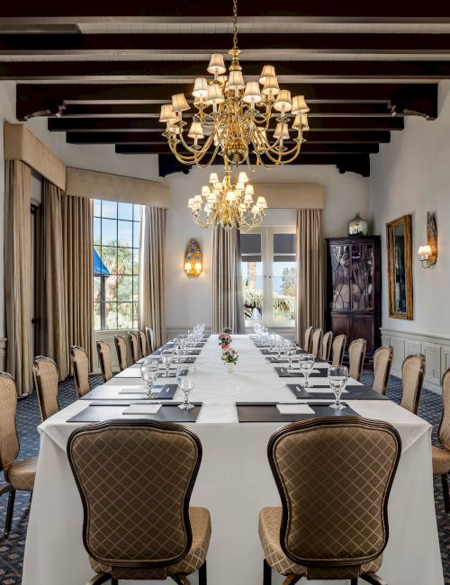 An elegant banquet hall with a long set table, cushioned chairs, and ornate gold chandeliers hanging overhead.