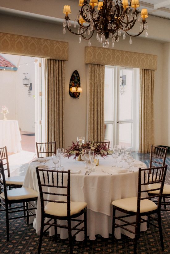 A round table set for a formal dinner with white tablecloths, elegant place settings, floral centerpiece, and black-chairs in a bright, chandelier-lit room.