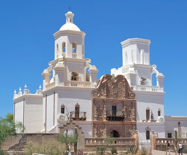 A grand white church with two tall towers, ornate brown central doorway, blue sky, and surrounding dry landscape. Ending with a period.