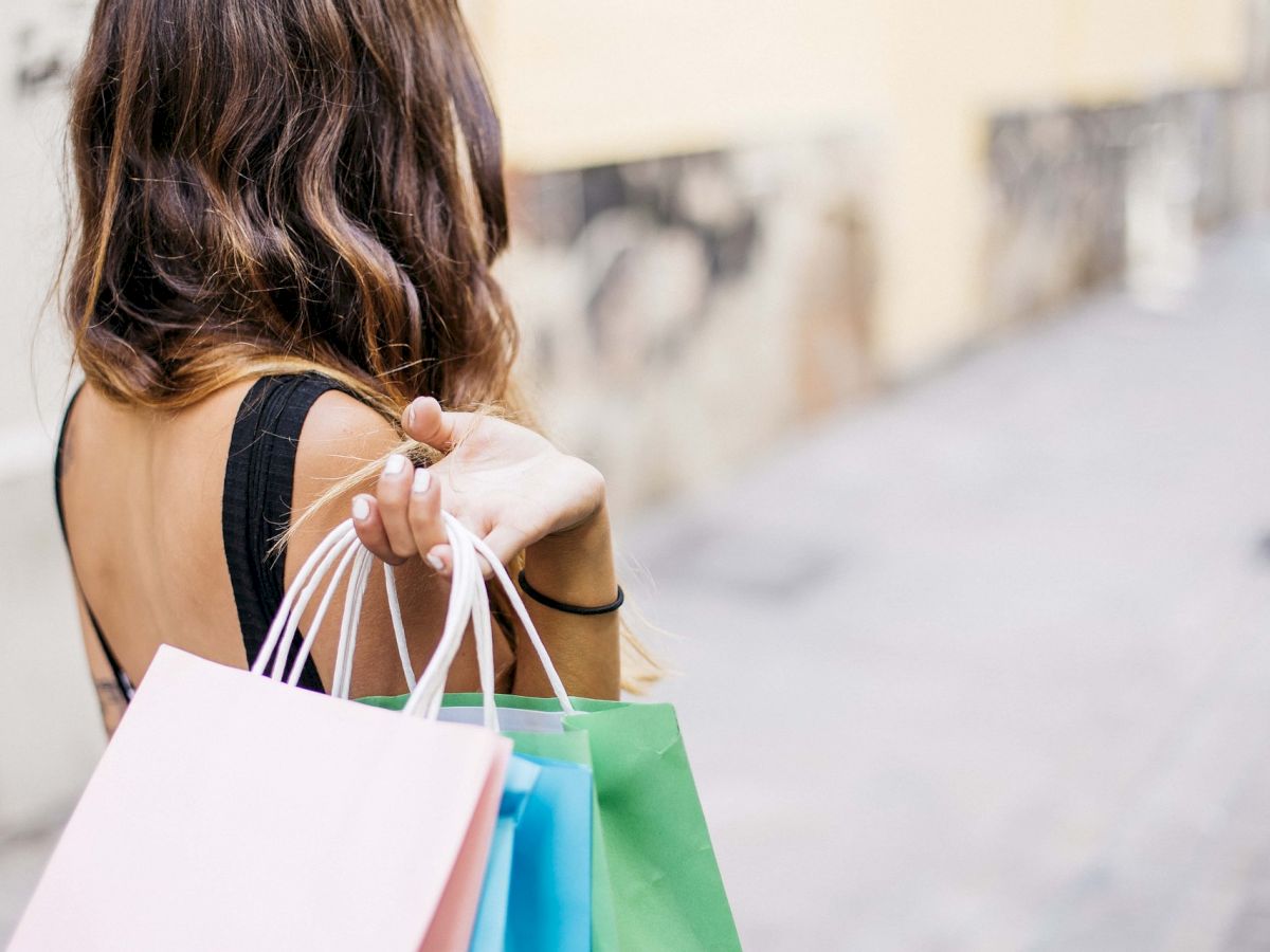 A person with wavy hair holds several colorful shopping bags, seen from behind, walking down a sunny street.